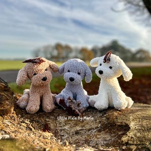 May include: Three crocheted plush dog toys in different colors: brown, gray, and white. The toys are sitting on a tree trunk with a blurred background of trees and sky.
