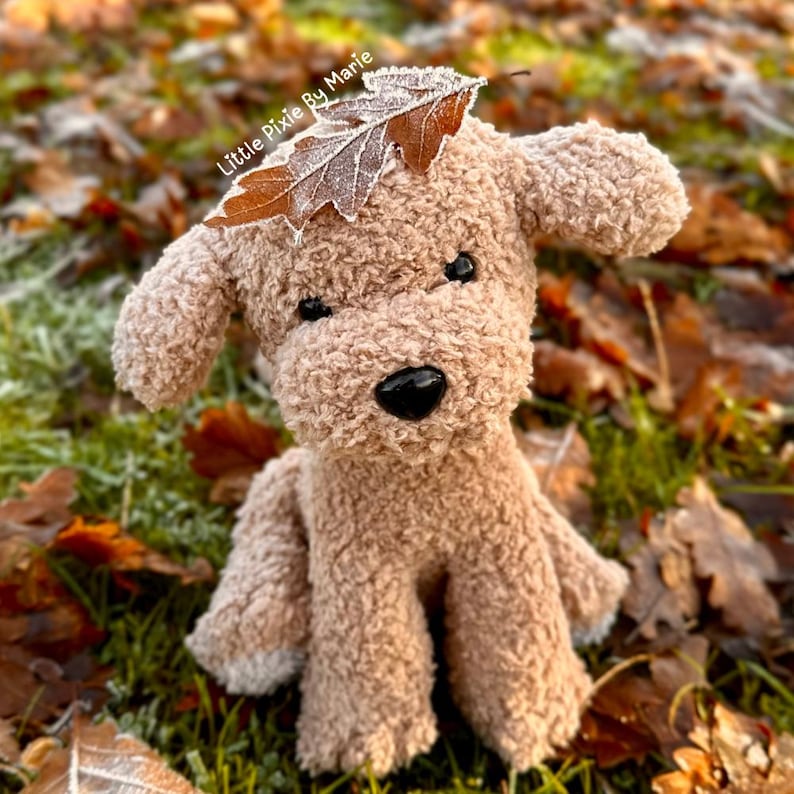 May include: A light brown plush toy dog with a black nose and eyes, sitting on a bed of brown leaves. The dog is wearing a brown leaf on its head. The text "Little Pixie By Marie" is visible in the image.