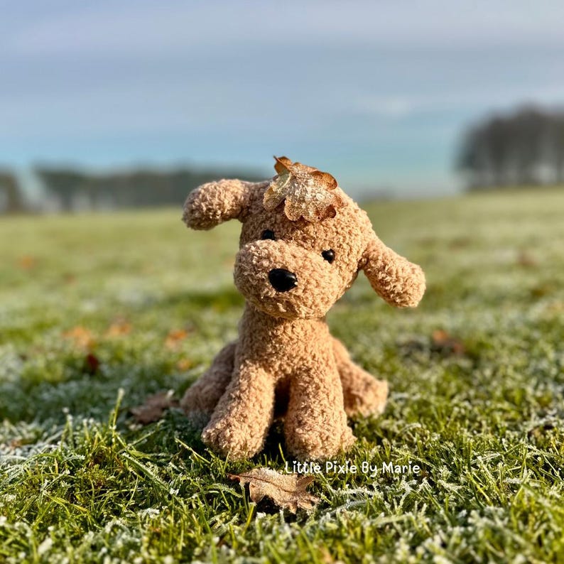 May include: A brown crocheted stuffed dog toy with a leaf on its head, sitting on a grassy surface covered in frost.