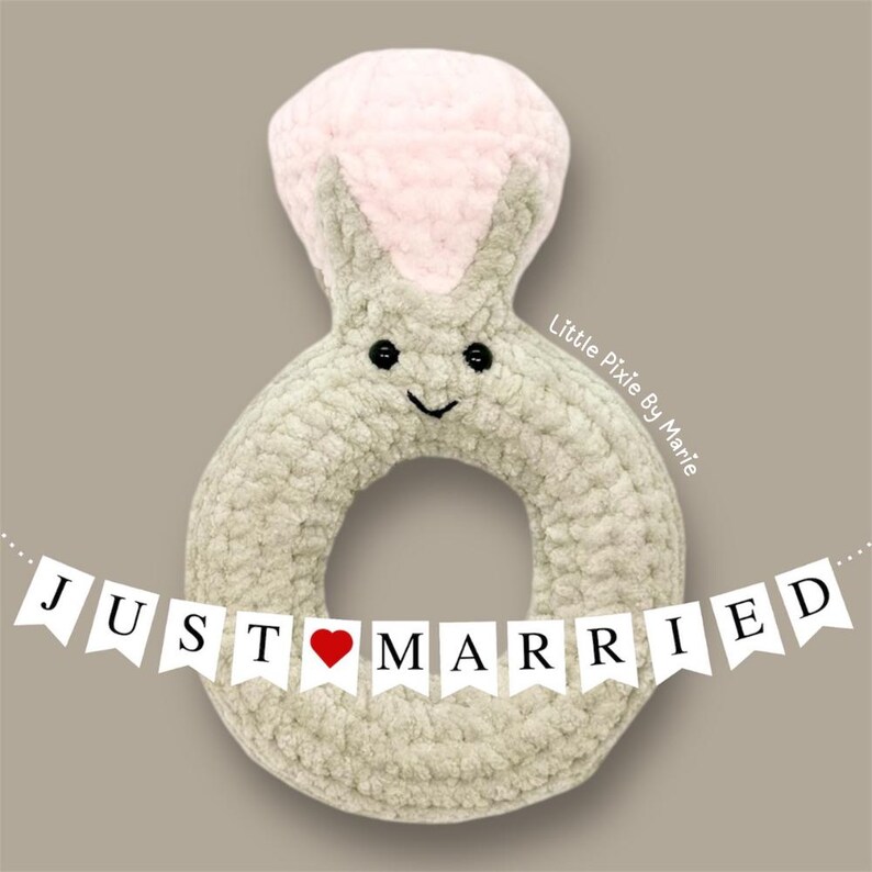 May include: A crocheted ring-shaped toy with a pink top and a smiling face. The toy is decorated with a banner that reads "JUST MARRIED" with a red heart.