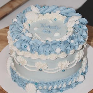 May include: A two-tiered light blue cake decorated with white frosting and seashells. The top tier has the words "Twenty One" written in white frosting. The cake is decorated with blue and white frosting swirls and white seashells.