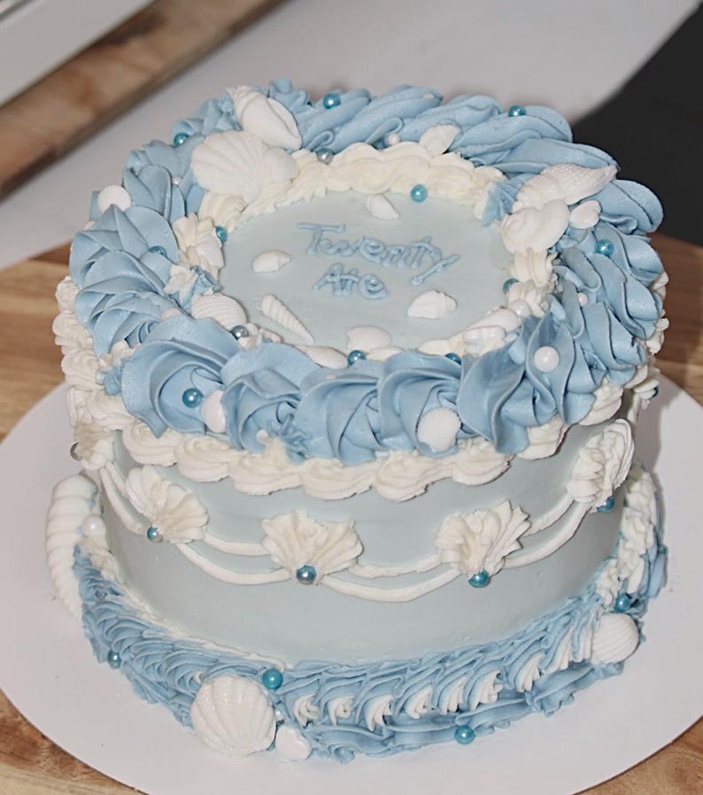 May include: A two-tiered light blue cake decorated with white frosting and seashells. The top tier has the words "Twenty One" written in white frosting. The cake is decorated with blue and white frosting swirls and white seashells.