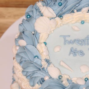 May include: A blue and white cake decorated with seashells and blue and white sprinkles. The cake says "Twenty Ate" on the top.