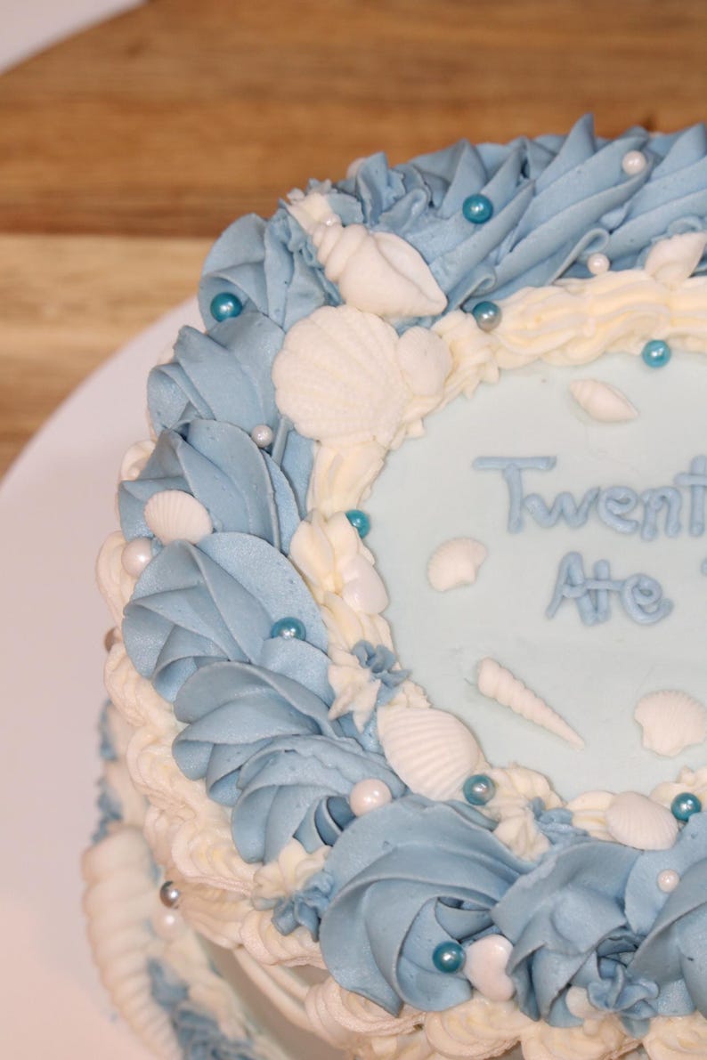 May include: A blue and white cake decorated with seashells and blue and white sprinkles. The cake says "Twenty Ate" on the top.