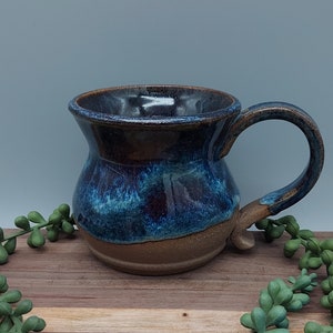 May include: A handmade ceramic mug with a blue and brown glaze. The mug has a wide, rounded base and a flared rim. The handle is attached to the side of the mug.