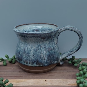 May include: A ceramic mug with a blue and white glaze. The mug has a wide base and a flared rim. The handle is attached to the side of the mug.