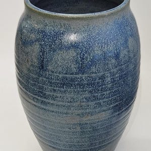 May include: A handcrafted ceramic vase with a textured, blue glaze. The vase has a wide, rounded body and a slightly flared rim. Horizontal lines encircle the vase, adding visual interest. The color palette includes shades of blue and gray.