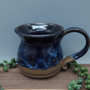 May include: A handmade ceramic mug with a dark blue and brown glaze. The mug has a wide, rounded body and a handle.