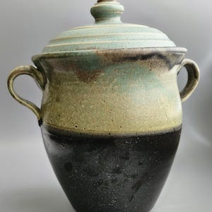 May include: A handcrafted ceramic jar with a lid. The jar features a two-handled design and a color gradient from black to olive green to a light blue-green on the lid. The jar is a decorative piece for the home.