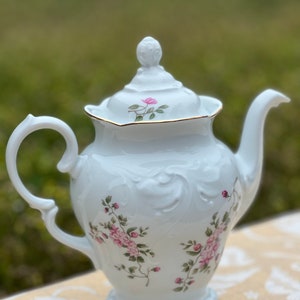 May include: White porcelain teapot with a floral design featuring pink roses and green leaves. The teapot has a gold trim around the lid and a fluted base.