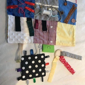 May include: A multi-coloured fabric activity mat with various textures and objects for babies and toddlers to explore. The mat features a variety of textures, including soft fabrics, ribbons, and buttons. There is a small black and white polka dot square with colourful ribbons attached. The mat also includes a wooden ring with a red and white fabric strip attached. The mat is labelled with the letter "C".