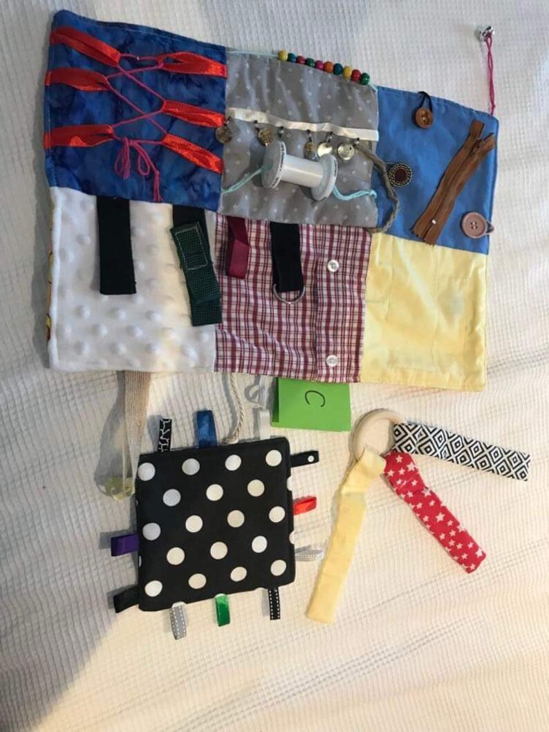 May include: A multi-coloured fabric activity mat with various textures and objects for babies and toddlers to explore. The mat features a variety of textures, including soft fabrics, ribbons, and buttons. There is a small black and white polka dot square with colourful ribbons attached. The mat also includes a wooden ring with a red and white fabric strip attached. The mat is labelled with the letter "C".
