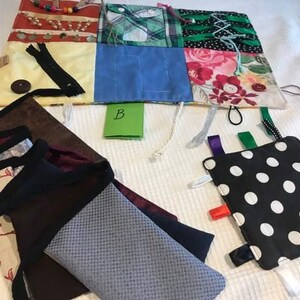 May include: A multi-coloured fabric square with various textures and patterns, including a zipper, buttons, and ribbons. The square is surrounded by a white background.