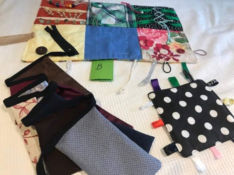 May include: A multi-coloured fabric square with various textures and patterns, including a zipper, buttons, and ribbons. The square is surrounded by a white background.