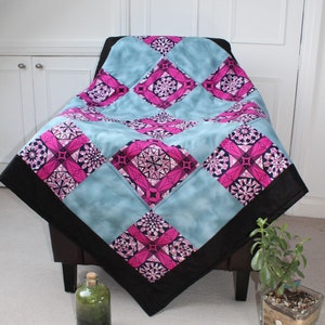 May include: A quilt with a geometric pattern in shades of pink, blue, and black. The quilt is draped over a chair with a black frame. A small potted plant is in the foreground.