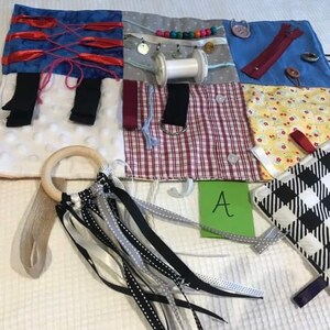 May include: A multi-coloured fabric activity mat with various textures and features for babies and toddlers. It includes a wooden ring, ribbons, a zipper, buttons, and a small fabric square with a black and white chequered pattern. The mat is labelled with the letter "A".