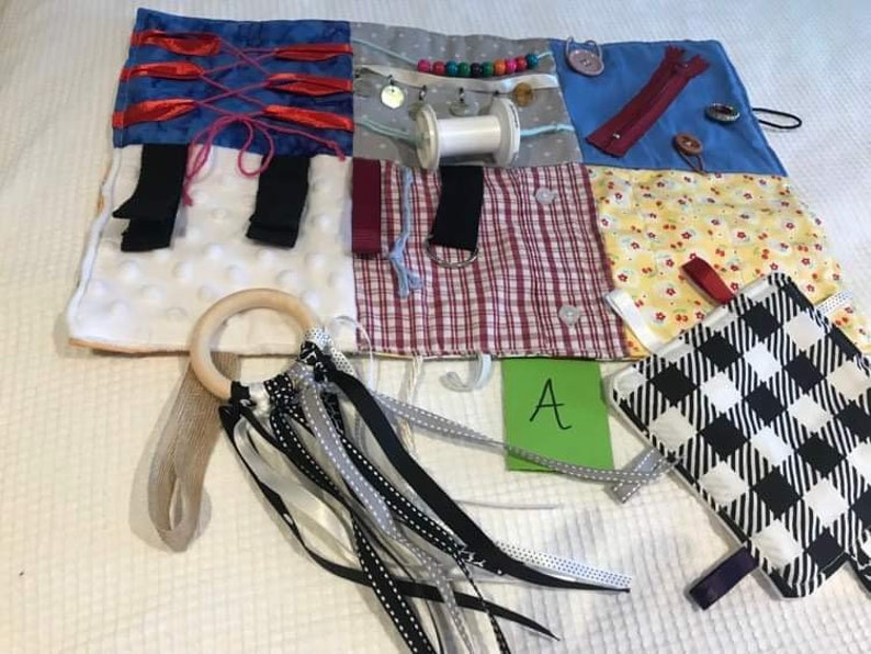 May include: A multi-coloured fabric activity mat with various textures and features for babies and toddlers. It includes a wooden ring, ribbons, a zipper, buttons, and a small fabric square with a black and white chequered pattern. The mat is labelled with the letter "A".