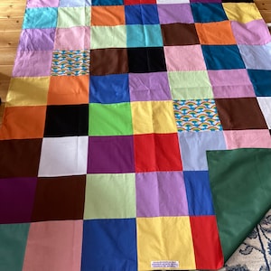 May include: A colourful patchwork quilt featuring squares of various colours such as blue, orange, purple, and green. Some squares have a rainbow pattern. The quilt is displayed on a wooden floor and a patterned rug.