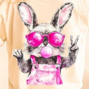 May include: A beige t-shirt with a cartoon bunny wearing pink sunglasses and a pink bubble. The bunny is wearing pink overalls and has a peace sign with its right hand.