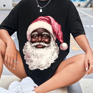 May include: Black t-shirt featuring a detailed illustration of Santa Claus with a white beard and a red hat with a white trim. The Santa Claus illustration has a dark skin tone. The t-shirt is a casual, oversized style.