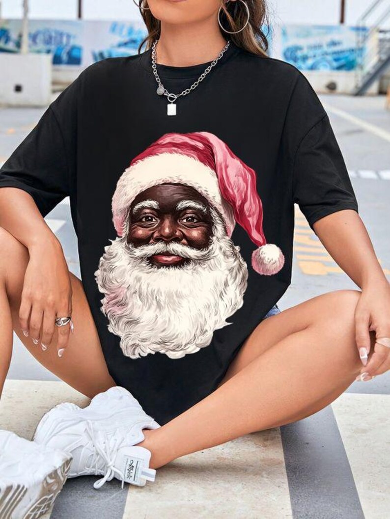 May include: Black t-shirt featuring a detailed illustration of Santa Claus with a white beard and a red hat with a white trim. The Santa Claus illustration has a dark skin tone. The t-shirt is a casual, oversized style.