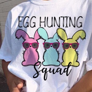Puede incluir: Camiseta blanca con un gráfico de tres conejos de Pascua con gafas de sol. Los conejos son rosa, azul y amarillo. El texto "Egg Hunting Squad" está impreso en la camiseta.
