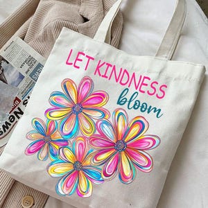 K&ouml;nnte beinhalten: Wei&szlig;e Stofftasche mit bunten Blumen und dem Text "LET KINDNESS bloom".