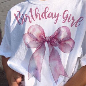 Może przedstawiać: Biała koszulka z napisem "Birthday Girl" w r&oacute;żowym kolorze i r&oacute;żową wstążką w stylu akwareli.