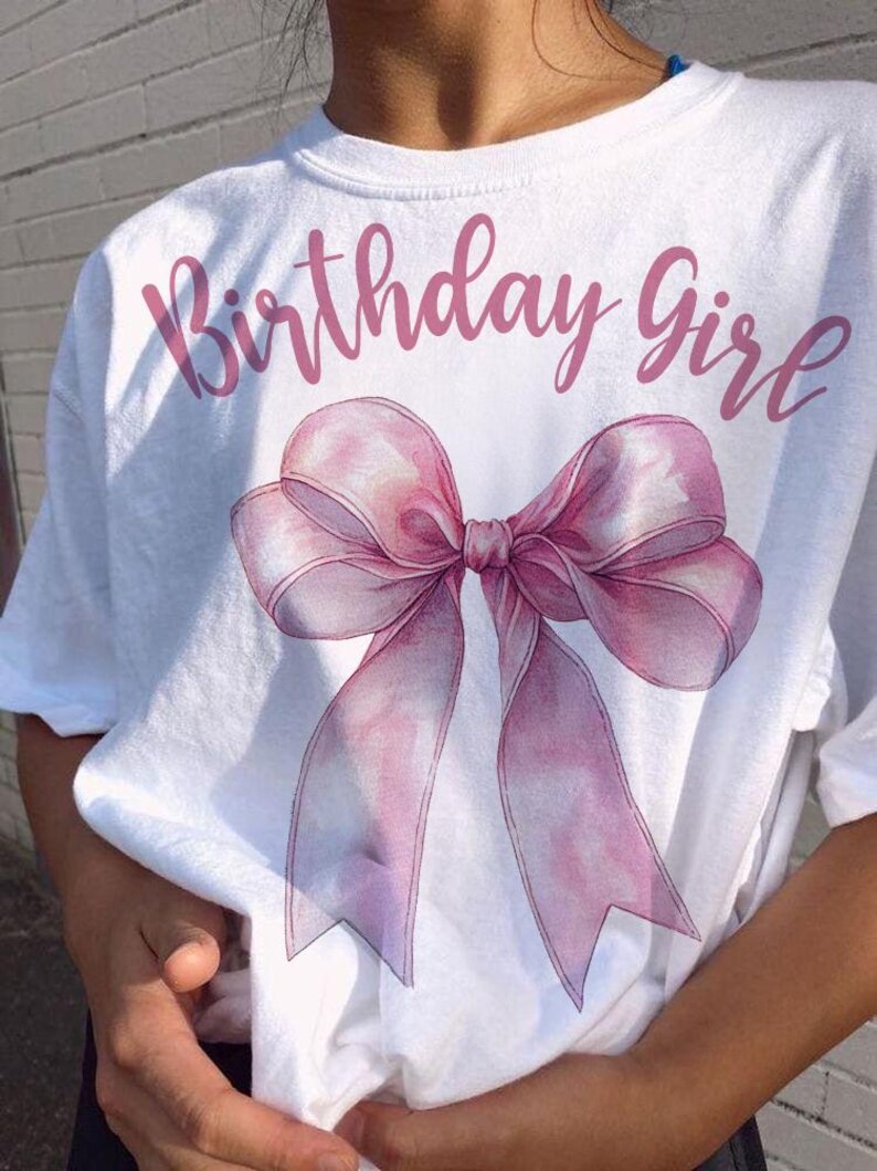 Może przedstawiać: Biała koszulka z napisem "Birthday Girl" w r&oacute;żowym kolorze i r&oacute;żową wstążką w stylu akwareli.