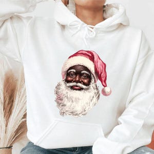 May include: White hooded sweatshirt featuring a graphic of Santa Claus with a dark complexion. Santa is wearing a red and pink hat and has a white beard. The design is centered on the front of the sweatshirt.