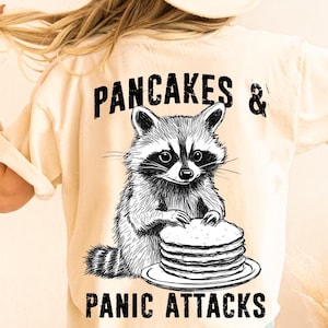 May include: A cream coloured t-shirt with a black and white illustration of a raccoon holding a stack of pancakes. The text "PANCAKES & PANIC ATTACKS" is printed on the shirt.