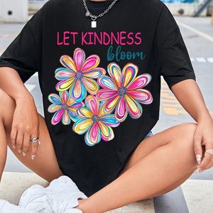 K&ouml;nnte beinhalten: Schwarzes Oversize-T-Shirt mit dem Text "Let Kindness Bloom" und einem bunten Blumenmuster.