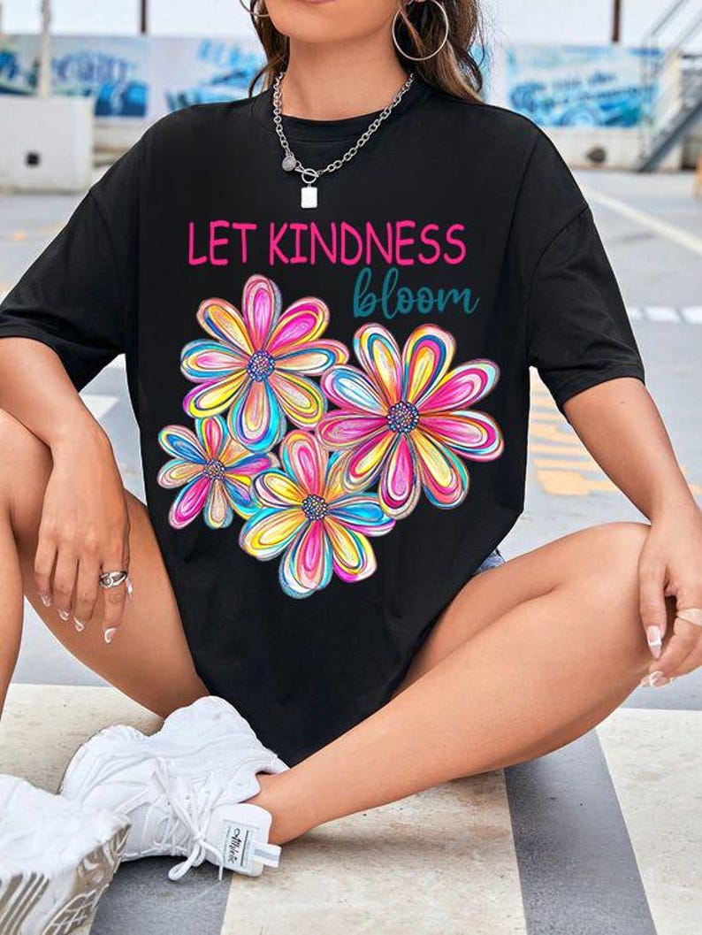 K&ouml;nnte beinhalten: Schwarzes Oversize-T-Shirt mit dem Text "Let Kindness Bloom" und einem bunten Blumenmuster.