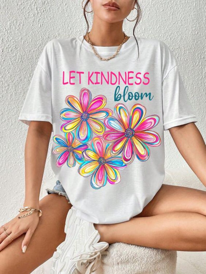 K&ouml;nnte beinhalten: Wei&szlig;es T-Shirt mit einem grafischen Druck von bunten Blumen und dem Text "Let Kindness Bloom".