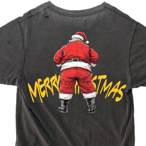 Può includere: Una maglietta grigia scura con una grafica gialla di Babbo Natale con il testo "Merry Christmas" sul retro.