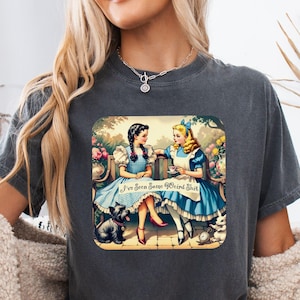 May include: A charcoal gray short-sleeve t-shirt featuring a graphic print of two characters in blue dresses sitting on a bench with a small dog and the text I've Seen Some Weird Shit. The design includes floral elements and a vintage aesthetic.
