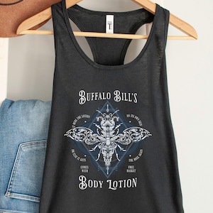 May include: A dark gray racerback tank top with the text "Buffalo Bill's Body Lotion" and a detailed insect graphic. The design includes a diamond shape with additional text. A brown hat and folded blue jeans are visible in the background.