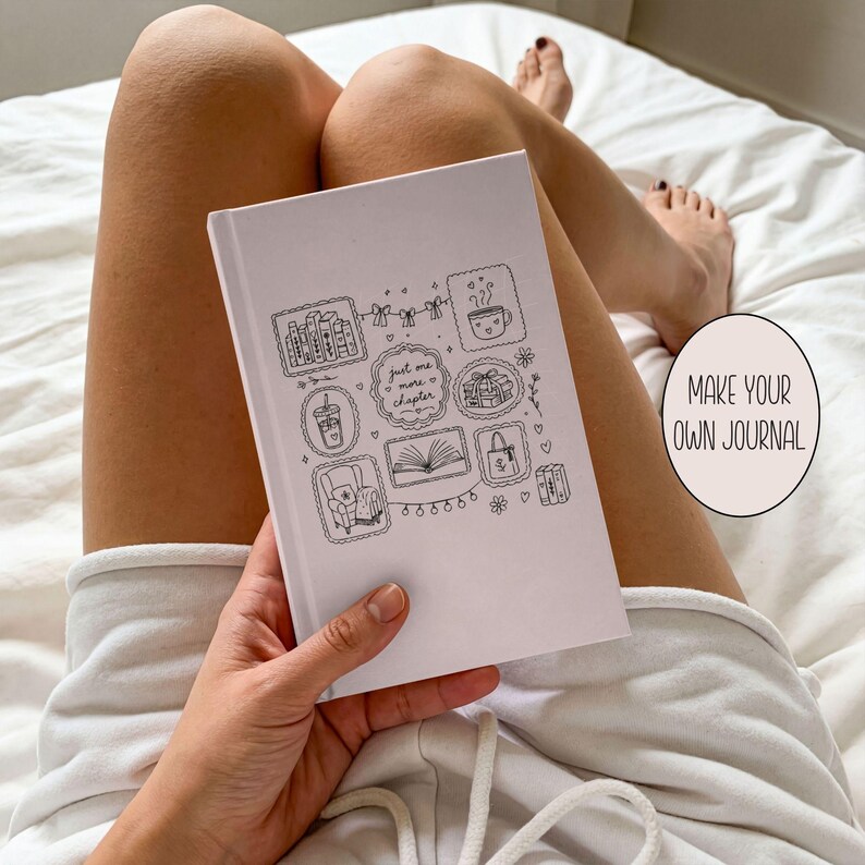 May include: A light pink journal featuring black line art illustrations of books, a coffee mug, a chair, and a tote bag, with the central text "just one more chapter" enclosed in a decorative frame, held against a white fabric background.