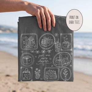 May include: A dark gray t-shirt featuring white line art illustrations of books, a coffee cup, a cozy armchair, and a tote bag. The text reads "just one more chapter" and "books tea cozy" along with "fairy tales, poems, little stories, old books."