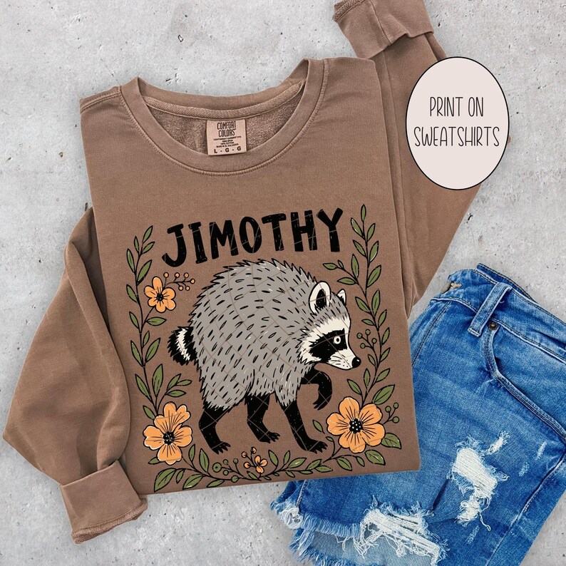 May include: A brown long-sleeve sweatshirt featuring a graphic of a raccoon surrounded by orange flowers and green leaves. The text "JIMOTHY" is printed in black above the raccoon. A circular white label reads "PRINT ON SWEATSHIRTS".