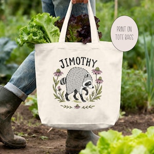 May include: A cream-colored canvas tote bag featuring a graphic illustration of a gray raccoon surrounded by pink flowers and green leaves. The text "JIMOTHY" is printed in black above the raccoon. The bag has two sturdy handles.