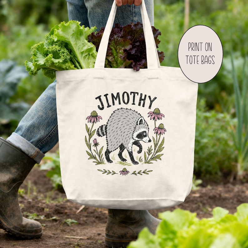 May include: A cream-colored canvas tote bag featuring a graphic illustration of a gray raccoon surrounded by pink flowers and green leaves. The text "JIMOTHY" is printed in black above the raccoon. The bag has two sturdy handles.