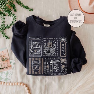 Op de afbeelding: Een donker marineblauw sweatshirt met ronde hals en een witte grafische print van een bibliotheekkaart met de tekst fully booked, een stapel boeken met een kaars, een bloemrijke boekenlegger, een koffiebeker en een open boek.