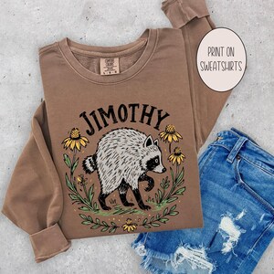 May include: A brown long-sleeve sweatshirt featuring a graphic of a raccoon surrounded by yellow flowers and green leaves. The text JIMOTHY is printed in black above the raccoon. A circular label on the right reads PRINT ON SWEATSHIRTS.
