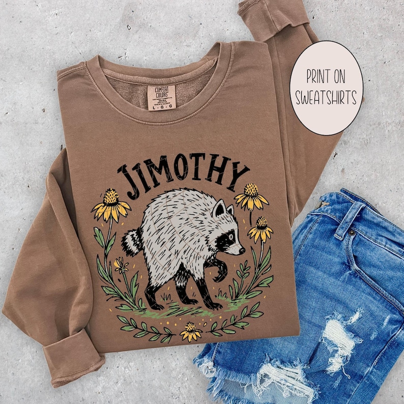 May include: A brown long-sleeve sweatshirt featuring a graphic of a raccoon surrounded by yellow flowers and green leaves. The text JIMOTHY is printed in black above the raccoon. A circular label on the right reads PRINT ON SWEATSHIRTS.