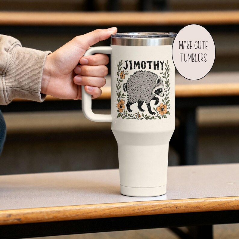May include: A cream-colored insulated tumbler with a handle and a lid, featuring a graphic of a gray raccoon surrounded by a floral wreath and the name JIMOTHY printed above. A circular text overlay on the right reads MAKE CUTE TUMBLERS.