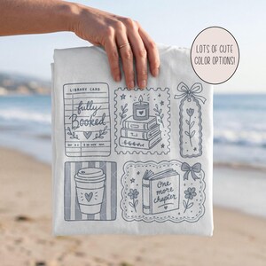 Op de afbeelding: Een wit t-shirt met een grijze grafische print van illustraties in boekenthema, waaronder een bibliotheekkaart met de tekst fully booked, een stapel boeken met een kaars, een bloemenbladwijzer, een koffiebeker en een open boek met one more chapter.