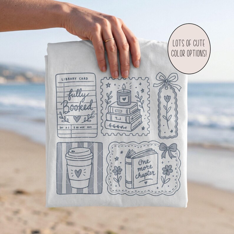 Op de afbeelding: Een wit t-shirt met een grijze grafische print van illustraties in boekenthema, waaronder een bibliotheekkaart met de tekst fully booked, een stapel boeken met een kaars, een bloemenbladwijzer, een koffiebeker en een open boek met one more chapter.
