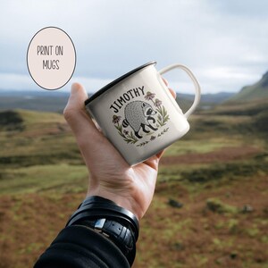 May include: A white enamel mug with a black rim featuring a graphic illustration of a raccoon surrounded by flowers and the text "JIMOTHY" arched above the animal. The mug is held against a blurred outdoor landscape background.