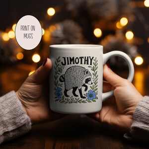 May include: A white ceramic mug featuring a black and white illustration of a raccoon surrounded by green leaves and blue flowers. The name JIMOTHY is printed in black capital letters above the raccoon. The mug has a curved handle on the right side.